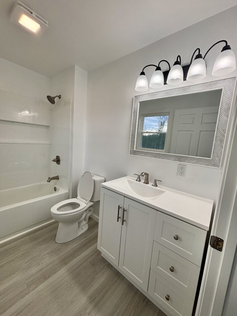 Updated bathroom with white vanity, toilet, mirror, and overhead lighting.